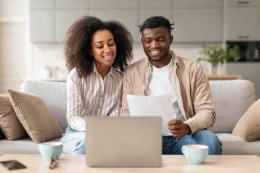 Mutlu siyah milenyum eşleri faturalarını dizüstü bilgisayarla ödüyor, kanepede oturuyor ve ev içinde dizüstü bilgisayar kullanıyor, çiftler internet bankacılığı kullanarak mülk satın alıyor.