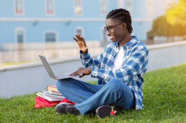 Siyah adam el sallayarak diz üstü bilgisayara el sallıyor, uzaktan dijital dersten zevk alıyor, dışarıda kampüs parkında oturuyor. Öğrenci uzak eğitimine bilgisayar ve internet üzerinden katılıyor