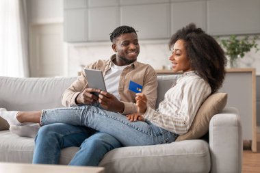 Çevrimiçi alışveriş başvurusu. Gülümseyen siyah çift dijital tablet ve kredi kartı kullanıyor, internet mağazasından internet alımı sipariş ediyor, banka hesaplarını kontrol ediyor, evde ödemeler yapıyor.