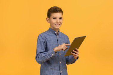 Mesafe öğrenme. Gülümseyen çocuk dijital tablet tutuyor ve kameraya bakıyor, mutlu erkek çocuk çevrimiçi çalışmak için modern teknolojileri kullanıyor, sarı stüdyonun arka planında duruyor.