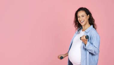 Koltuk değnekleriyle egzersiz yapan, kameraya bakan ve gülümseyen, fotokopi çeken gülümseyen hamile kadın. Pembe arka planda, halterler kullanarak kadın eğitimi almak sağlıklı bir beklenti. Hamilelik sırasında spor