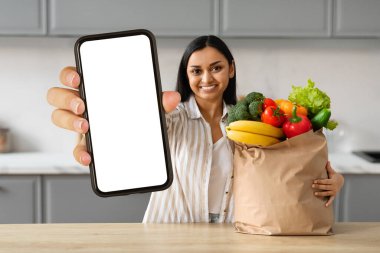 Gülümseyen genç Hintli kadın internetten alışveriş siparişi veriyor, mutfakta masanın yanında duruyor, elinde içi taze organik meyve ve sebzelerle dolu bir poşet tutuyor, telefonu beyaz ekranlı gösteriyor, mobil uygulama