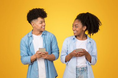 Neşeli Afro-Amerikan gençleri günlük giysiler içinde mutlu mesut birbirlerine bağlanıyorlar, ellerinde akıllı telefonlar, muhtemelen sosyal medya içeriklerini paylaşıyorlar, parlak sarı bir arka plana karşı.