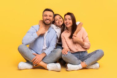 Sarı Stüdyo 'nun arka planına karşı bir arada oturan baba, anne ve küçük kızın mutlu Avrupalı aile fotoğrafı. Neşeli Kız, Kameraya Gülümseyen Ebeveynlere Kucaklıyor