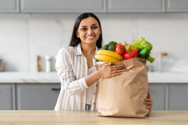 Evdeki modern gri mutfakta poz veren, kağıt torba dolusu taze eko meyve ve sebze dolu neşeli, genç Hintli kadın, alışveriş servisinin keyfini çıkarıyor, boş alan.