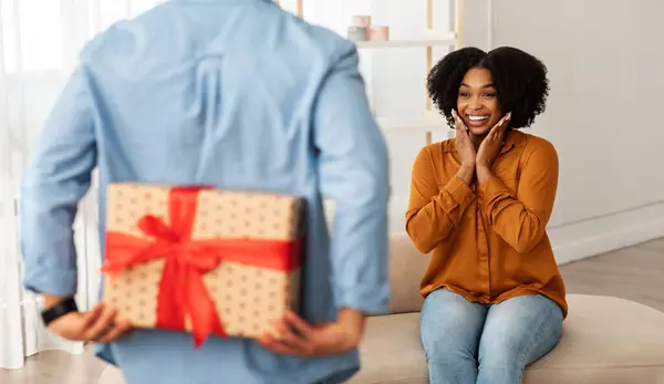 An excited woman with hands on cheeks sits on a sofa, looking with surprise at a person presenting her with a large gift wrapped in heart-patterned paper and a red ribbon