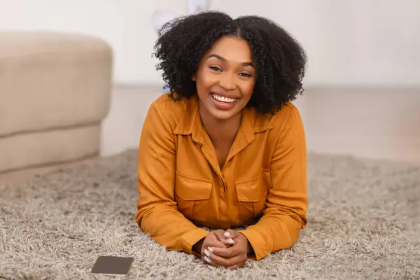 Cheerful pretty millennial curly african american woman lies on floor with phone, enjoy spare time in living room interior. Gadget and social networks at home, rest alone