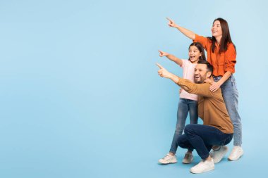 Şuraya bak. Küçük kızıyla birlikte mutlu beyaz bir aile, parmak kaldırıyor, Mavi Stüdyo Arkaplanı Üzerinde Mesaj İlanı Vermek İçin Boş Yer Gösteriyor. Ebeveynler ve Kız Çocuk Kopya Alanı Gösteriyor