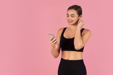 Gülümseyen genç sporcu, akıllı telefonuna bakarken kablosuz kulaklıklarını ayarlıyor, güzel atletik bayan müzik dinliyor, gündelik ortamlarda teknolojinin tadını çıkarıyor, pembe arka planda duruyor.