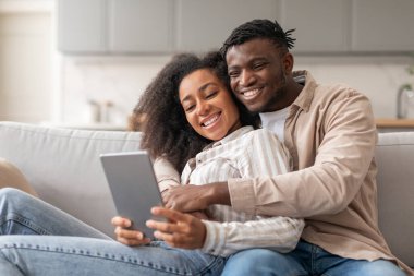 Afro-Amerikalı genç erkek ve kadın rahat modern oturma odasındaki tablet bilgisayarda internette geziniyor, kanepede dinleniyor ve hafta sonları rahat teknoloji zamanı geçiriyorlar. Web sitesi reklamı