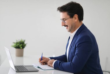 Masada dizüstü bilgisayarı olan neşeli beyaz iş adamı, modern beyaz ofis iç mimarisinde not defteri, profil. Planlama işi, iş, başlangıç için fikir yaratmak