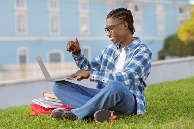 Siyah adam diz üstü bilgisayar kamerasına el sallıyor, uzaktan dijital dersi onaylıyor, kampüs parkında çimlerde oturuyor. Öğrenci şu şekilde gösteren bir eğitim web sitesi öneriyor