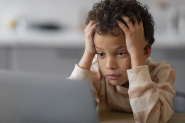 Evde eğitim zorluğu. Evde dizüstü bilgisayar kullanan ve kafaya dokunan üzgün siyah çocuk, ev ödevi yapmaktan yorulmuş çocuk, bilgisayar başında oturan depresyondaki çocuk, boş alan.