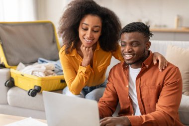 Keyifli Afro-Amerikan eşleri tatil planlarken dizüstü bilgisayar kullanıyorlar, biletleri ve oteli internetten ayırtıyorlar, bavulları boşaltılmış halde evde oturuyorlar, web gezisi gezileri için hazırlanıyorlar.