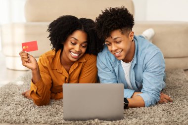 Neşeli genç Afro-Amerikalı erkek ve kadın alışverişkolikler, dizüstü bilgisayar, kredi kartı kullanırlar, siparişin keyfini çıkarırlar, oturma odasında yerde yatarlar. İnternetten satış, evden alışveriş.