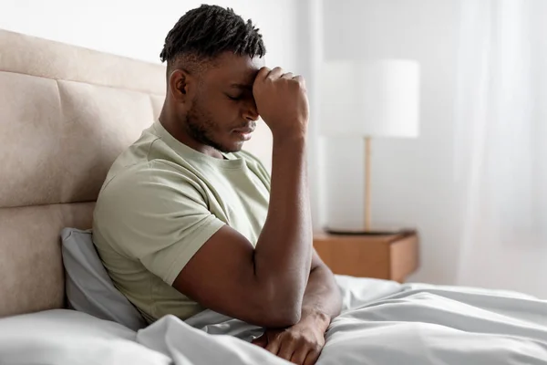 Unhappy black young man in bed pressing fist to forehead suffering from depression and chronic headache problem, waking up in the morning in modern bedroom at home, side view