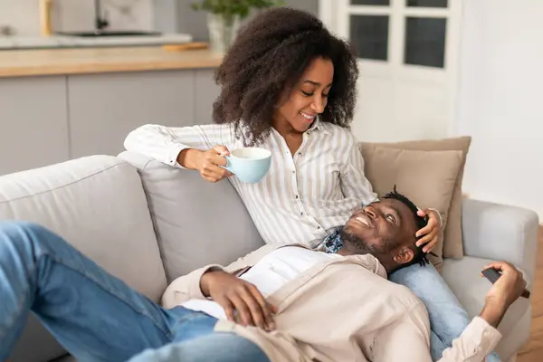 Happy Black Spouses Drinking Coffee Relaxing Together On Sofa, Watching TV In Comfortable Living Room, Man Resting Head On Wifes Legs, Enjoying Lazy Weekend Indoor