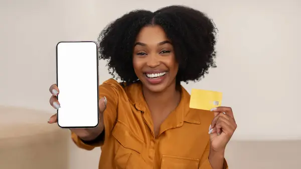 Smiling millennial african american curly woman show credit card and smartphone with empty screen in living room interior, panorama. Online shopping recommendation, sale, banking at home
