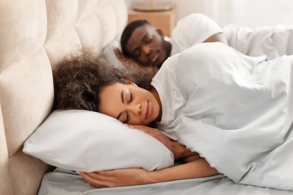African american young lady hugging pillow sleeping next to her husband, napping together in their bedroom, lying peacefully with eyes closed in the morning. Selective focus