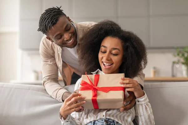 Smiling African American man surprising his woman with wrapped present in their cozy living room, giving St Valentines Day gift to wife, celebrating romantic holiday or relationship anniversary
