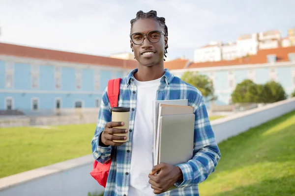 Portrait Of Happy Successful Black Student Guy Holding Laptop And Cup Of Takeaway Coffee Outdoor, Standing On Backdrop Of University Building, Smiling To Camera Carrying His Workbooks