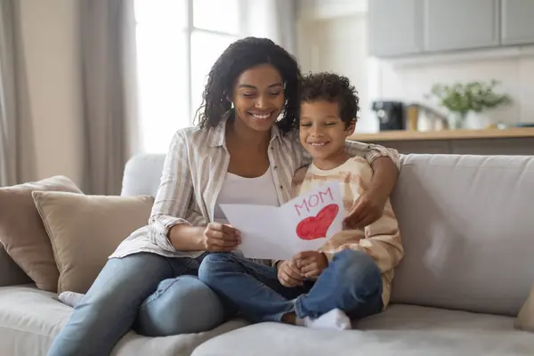 Mothers Day. Black Little Son Giving Greeting Card To His Happy Mom At Home, Loving Male Child Congratulating Mommy With Holiday While Sitting Together On Couch In Living Room, Free Space