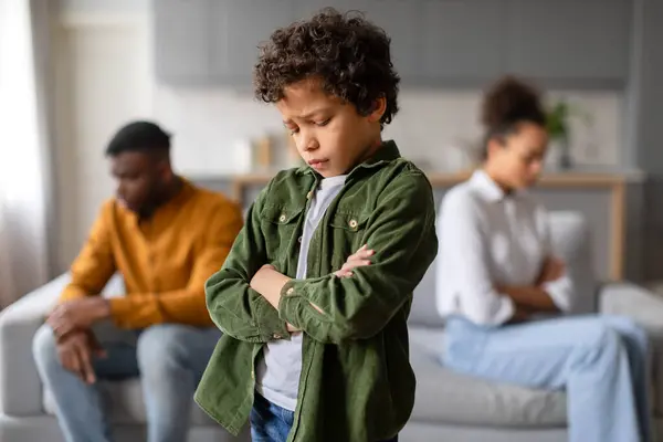 硫磺色的黑人男孩交叉着双臂站在前排 他的不安的表情与分别坐在后面的模糊的父母形成了鲜明的对比 — 图库照片