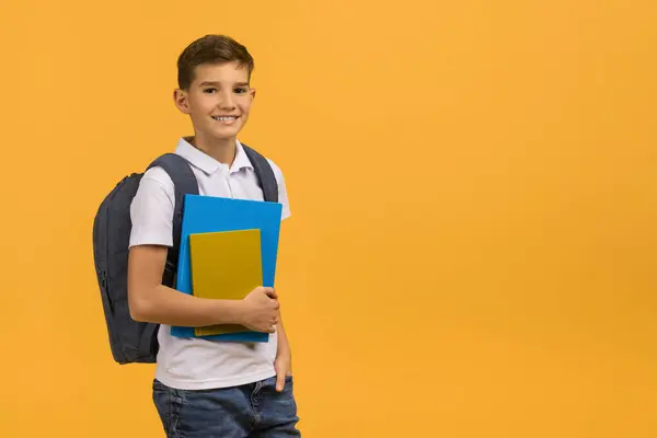 黄色いスタジオの背景に立ってバックパックと教科書を運ぶ笑顔の学校ボーイ 新しい学校の日の準備を描いた白いポロシャツとジーンズを着て陽気な男性の子供 コピースペース — ストック写真