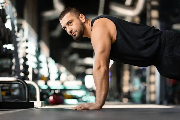 Motivated young male doing floor push-ups exercises while training in gym, athletic millennial man showcasing strength and determination, exercising in contemporary sport club interior, side view