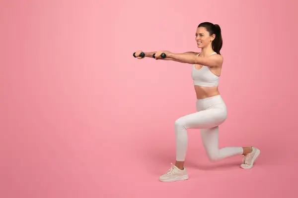 Happy strong millennial caucasian woman in white sportswear, squats with dumbbells, enjoy training, active lifestyle, isolated on pink background. Health care, sport and weight loss