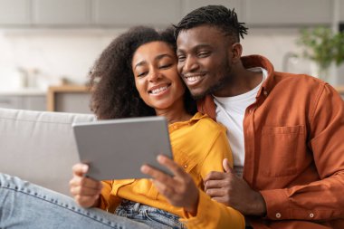 Neşeli Afro-Amerikalı eşler tablet bilgisayarla internette sörf yapıyor, çevrimiçi güzel filmlerin tadını çıkarırken sarılıyorlar, modern oturma odasının içinde dinleniyorlar, kanepede yan yana oturuyorlar.