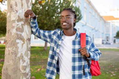 Sırt çantalı, ağaca yaslanmış üniversite kampüsünün dışında gülümseyen, başarılı bir genç öğrencinin portresi. Öğrencilik ve modern eğitim