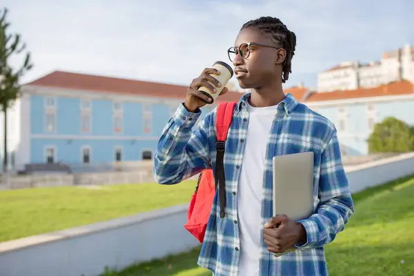 Black student guy with backpack and laptop drinking takeaway coffee looking away, enjoying break from education, standing on backdrop of modern university campus outdoors. Studentship lifestyle