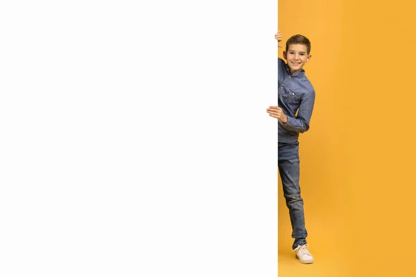 快乐的少年男孩 头戴粗斜纹棉布 从空白的白色垂直横幅后面露出微笑 快乐的少年男孩 站在鲜活的黄色背景下 展示广告的复制空间 — 图库照片
