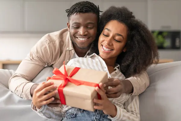 Romantic Present. Loving black husband presenting gift box to woman at home indoor, celebrating an anniversary of relationship together, hugging on sofa in living room. Valentines Day Offer