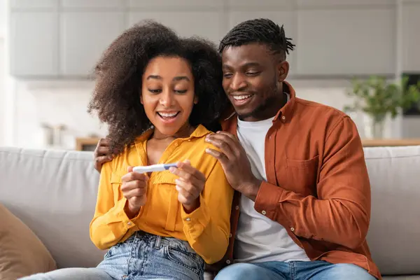 Well Be Parents. Joyful Married African American Couple Holding Positive Pregnancy Test, Sitting On Sofa Together, Celebrating And Hugging In Modern Living Room At Home. Family Moments, Childbirth