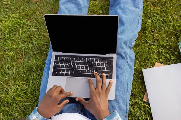 Unrecognizable black student guy studies websurfing on laptop with blank screen sitting on lawn outside, using computer for online education and work. E-learning offer. Mockup
