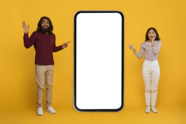 Harika bir online teklif, anlaşma, güzel mobil uygulama. Heyecanlı genç Hintli çift, reklam, el kol hareketi, sarı arka planda izole edilmiş beyaz ekranlı büyük telefonu işaret ediyor.
