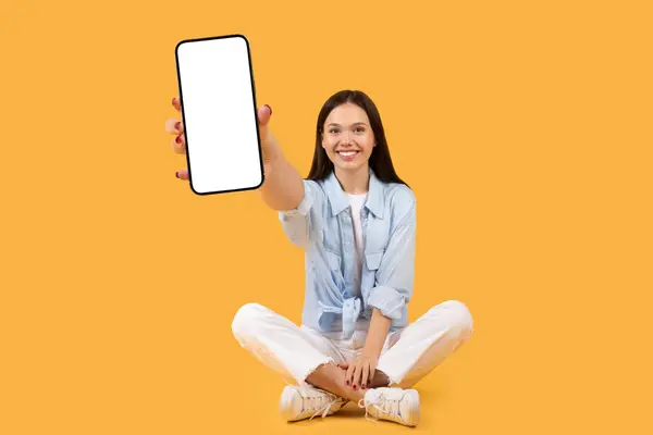 カメラに白いスマートフォンの画面を幸せに表示し 黄色の背景に座っているカジュアルな服装の陽気な若い女性 モックアップ — ストック写真