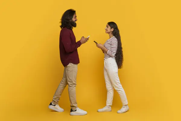 Dating mobile app, modern communication. Positive stylish millennials man and woman walking towards each other with smartphone in their hands, isolated on yellow studio background, full length