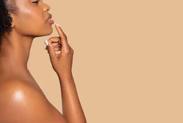 Contemplative young black woman in profile, gently touching her lips with finger, deep in thought, against soft beige background, free copy space
