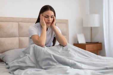 Genç bir kadın, elleri şakaklarında, yüzünde acı dolu bir ifadeyle yatakta oturuyor. Baş ağrısı ya da stres yaşadığını gösteriyor.