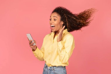 Canlı sarı bluzlu neşeli genç siyahi kadın pembe arka planda hareketli saçlarıyla akıllı telefonuna bakarken şaşkınlığını dile getiriyor.