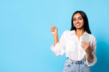 Güzel yeni uygulama. Gülümseyen Hintli genç bayan cep telefonu kullanıyor, harika mobil uygulama reklamları yapıyor, mavi stüdyo arka planında boş bir yeri işaret ediyor. Modern mobil iletişim