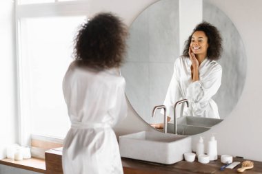 Güzellik Konsepti. Aynaya ve Dokunaklı Yüze Bakan Çekici Genç Zenci Kadın Modern Banyo 'da Cilt Bakımı yapan Beyaz Bornoz Giyen, Şımartan, Seçici Odaklanan Güzel Kadın