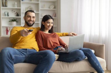 Evdeki koltukta dinlenen mutlu genç çift, dizüstü bilgisayar kullanan erkek ve kadın, mutlu milenyum eşleri oturma odasında birlikte vakit geçiriyorlar, ev işlerinin keyfini çıkarıyorlar, yakın çekim yapıyorlar.