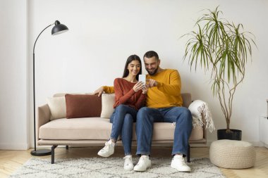 Evdeki koltukta dinlenirken cep telefonu kullanan güzel genç çift, modern akıllı telefonlu koltukta dinlenen mutlu beyaz eşler, yeni uygulama veya alışveriş sitelerine bakıyorlar,