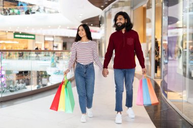 Alışveriş merkezinin önünden geçen, el ele tutuşan, gülümseyen renkli kağıt torbaları taşıyan, hafta sonları alışverişin keyfini çıkaran mutlu Hintli çift.