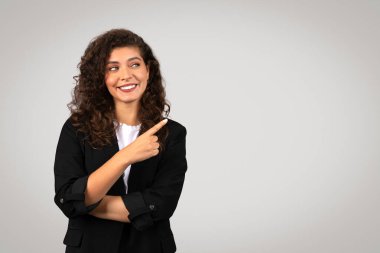 Siyah ceketli ışıl ışıl genç bir iş kadını, neşeli bir ifadeyle sağını işaret ediyor, yumuşak gri arka planda duruyor, iş afişi.