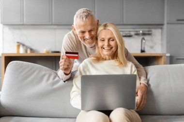 Heyecanlı üst düzey Avrupalı çift dizüstü bilgisayar ve kredi kartı kullanarak hafta sonu İnternet gezisi satın alıyor. Mutlu bir emekli adam ve kadın evdeki kanepede oturuyor, internetten mal ve hizmetleri ödüyor.